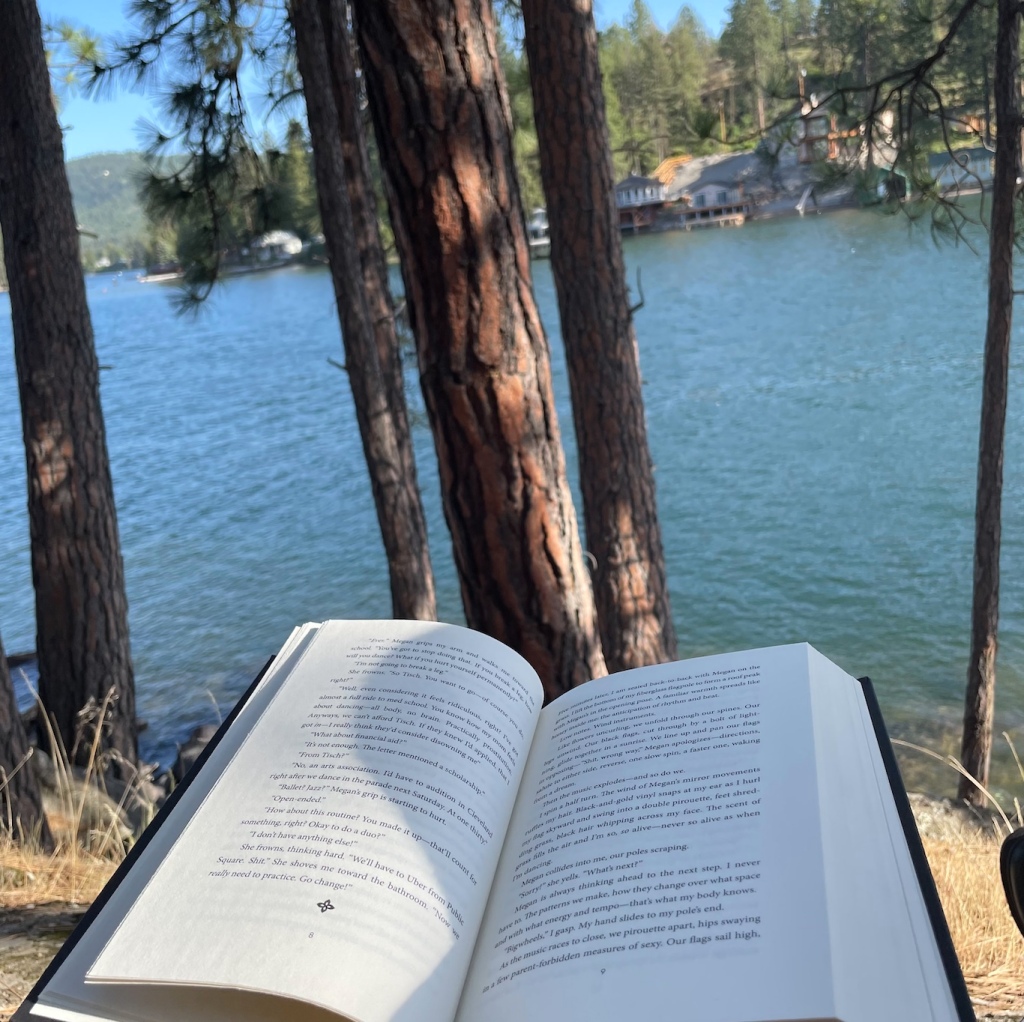 Book open to pages 8-9. A shadow covers most of the book. In the background there are 5 trees and the bank of a lake. In the way back of the picture is an unfocused house and dock.