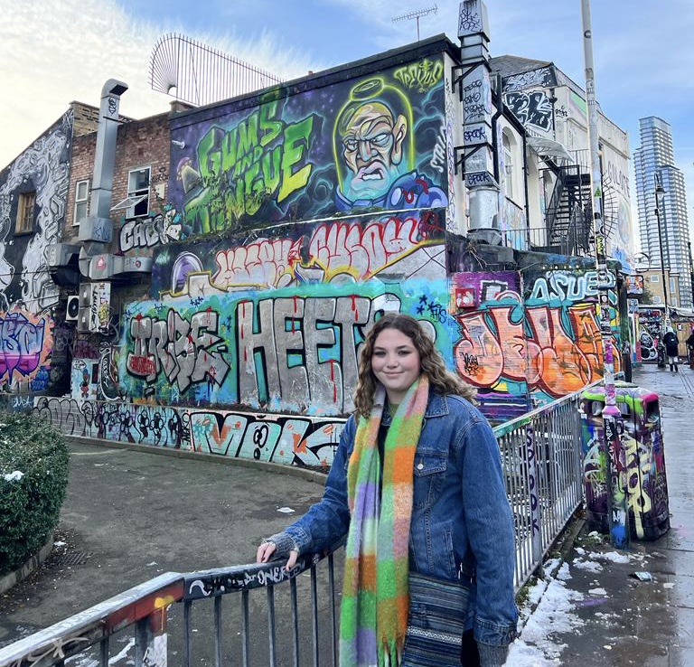 This image is of Olivia Andersen; she has brown hair and is wearing a green and orange scarf and a jean jacket. She is centered in front of a wall that has graffiti painted on the outside of the wall. 