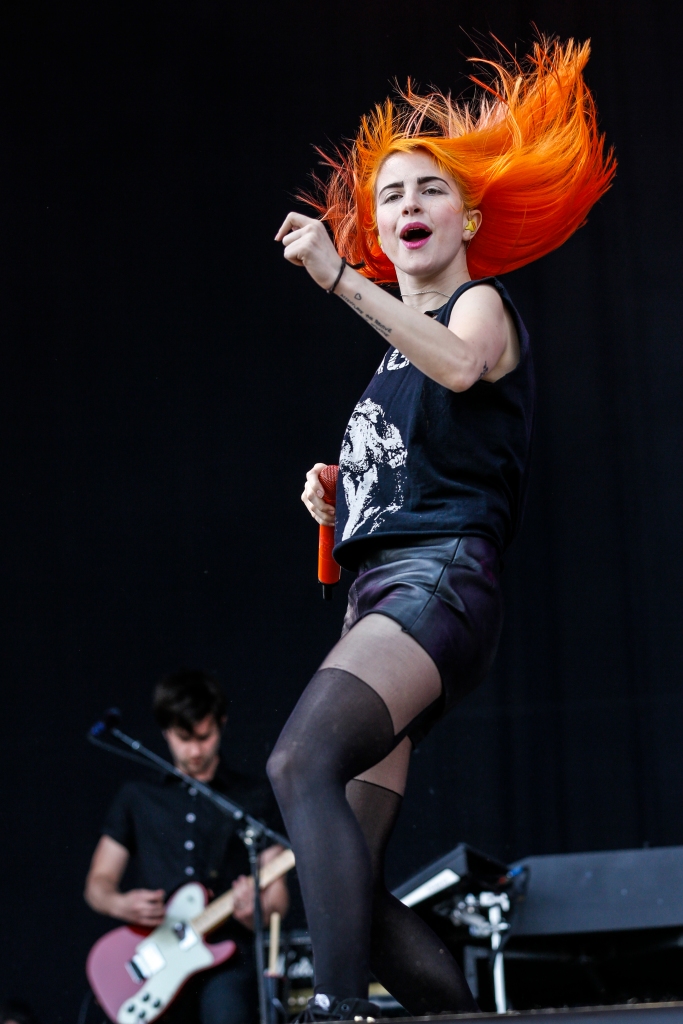 Picture of the lead singer in Paramore, Hayley Williams' singing to the audience. She has bright orange hair and is wearing black fishnets, a skirt, and a tank top. 