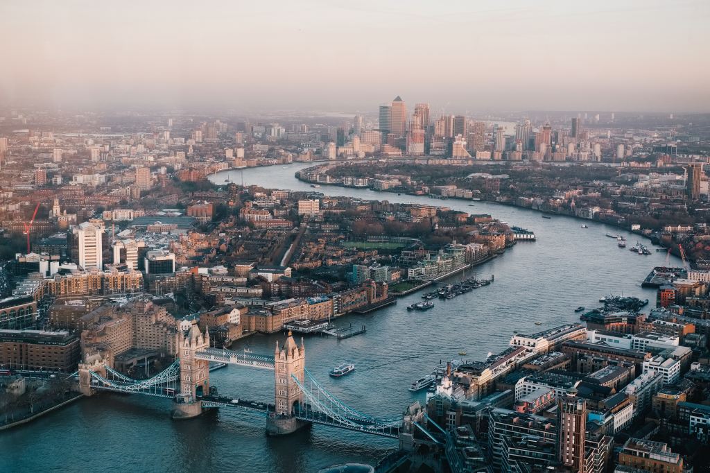 Photo depicts an aerial view of London, England during the day. At the top of the photo, the sky is mostly gray with a tinge of pink. The blue River Thames is flowing from the bottom left corner to the mid near top of the picture. Many boats and buildings can be seen. 