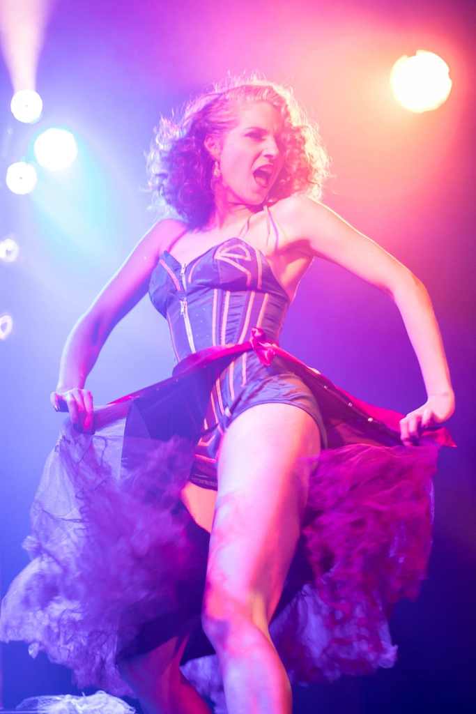 A young female-presenting burlesque performer wearing a tight black corset and translucent skirt flicks their skirt to reveal their bare thigh under orange and blue stage lighting.