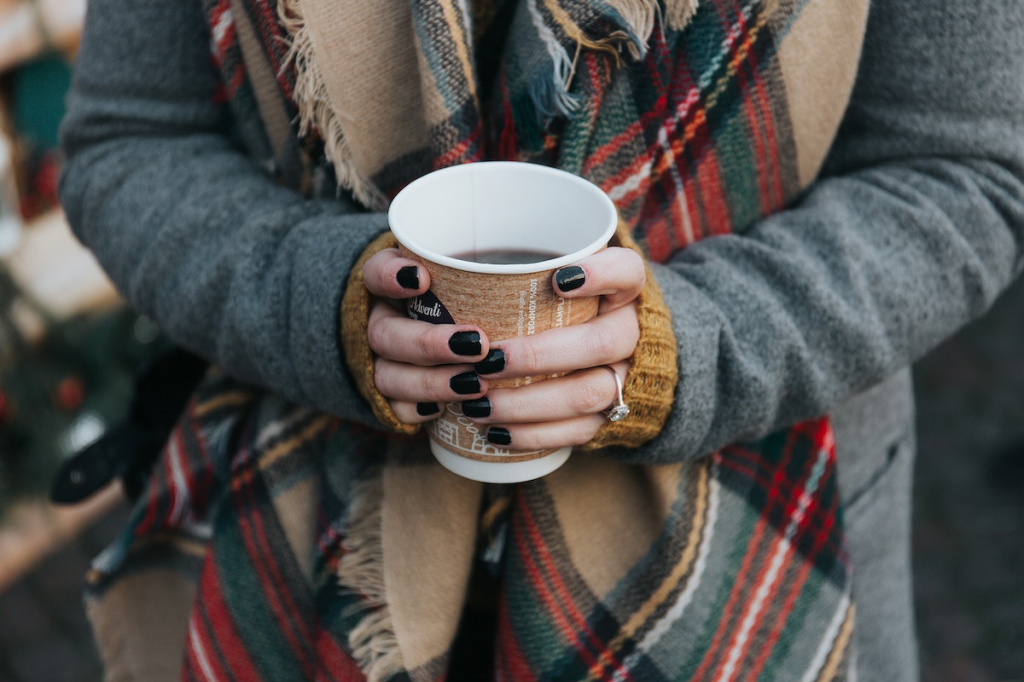 A women wears a grey coat with a flannel scarf. Her hands hold a paper coffee cup in front of her. 